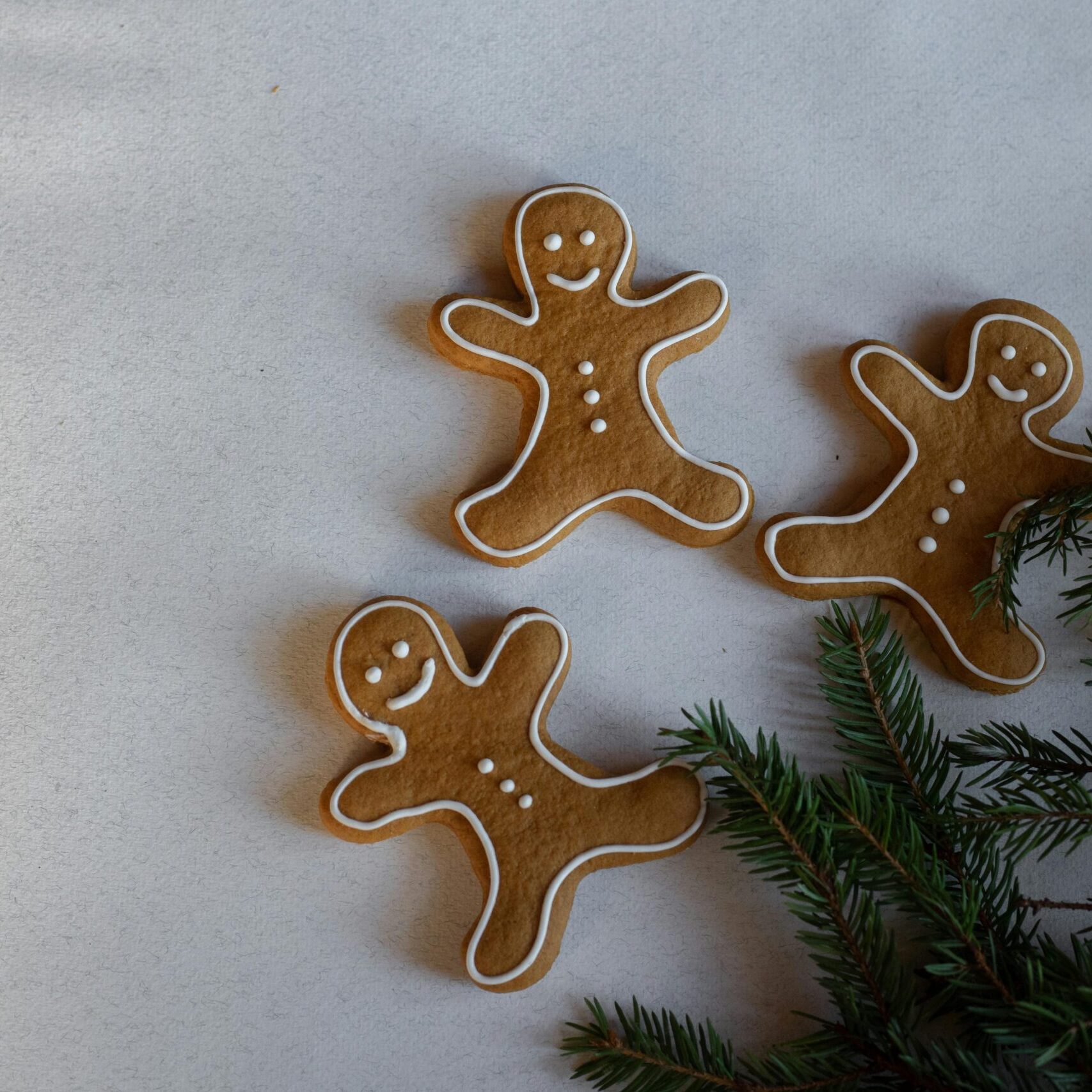 Three gingerbread men cookies with frosting on a surface next to pine leaves.